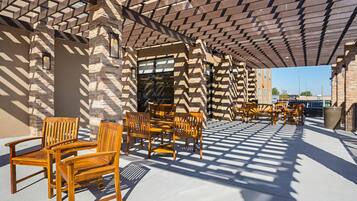 An outdoor seating area with wooden chairs and tables under a patterned roof, with a stone wall in the background.