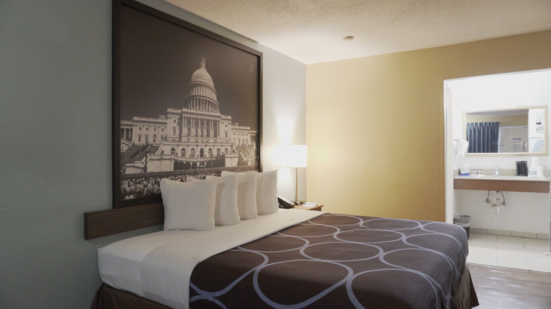 Habitación de hotel con una cama grande, una mesita de noche, una lámpara, un baño con lavamanos y espejo, y una pared con un cuadro de un edificio del capitolio.