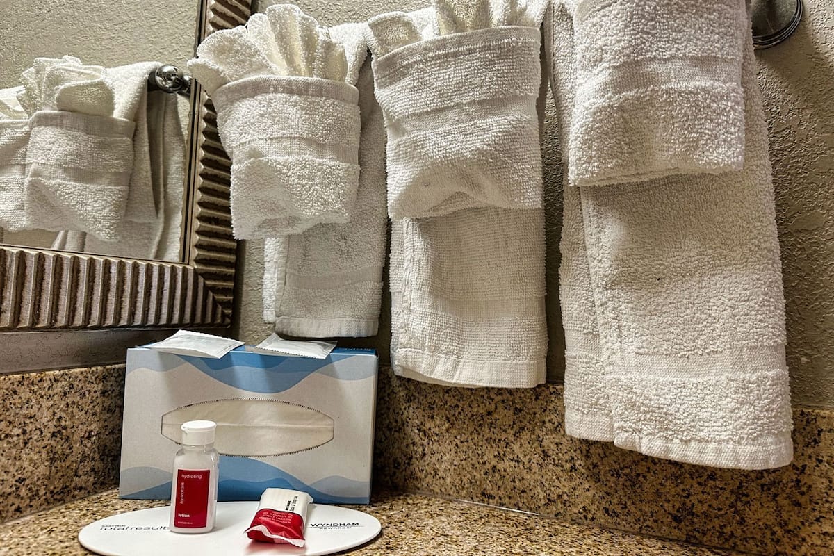 A bathroom with a mirror, a towel rack with multiple towels, a soap dispenser, a lotion bottle, and a box of toiletries.