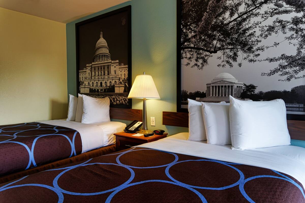 A hotel room with two beds, a nightstand with a lamp, a phone, and a wall painting of a building.