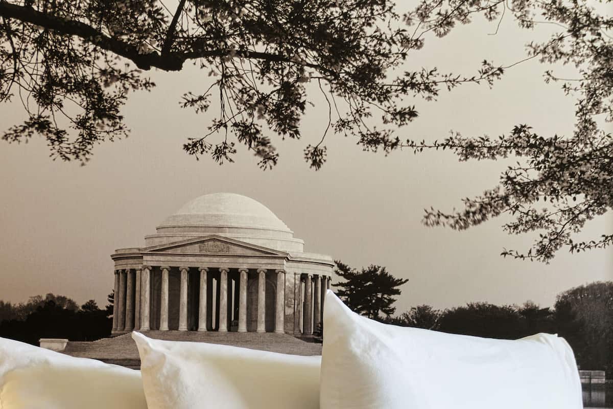 A bed with white pillows and a patterned tablecloth, with a classical building in the background.