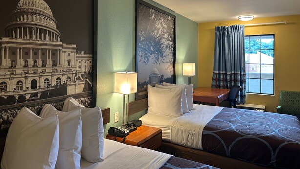 A hotel room with two beds, a nightstand, a lamp, a window with curtains, and a wall mural of the Capitol building.