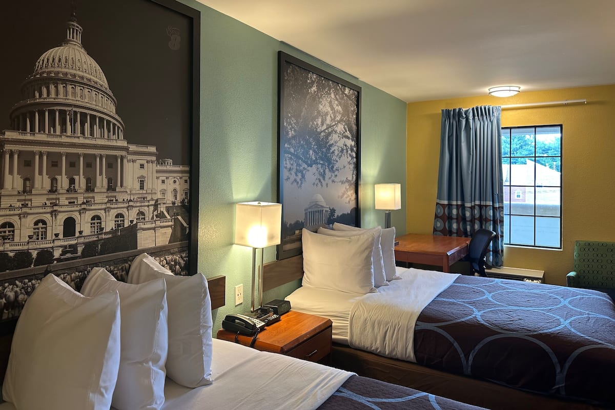 A hotel room with two beds, a nightstand, a lamp, a window with curtains, and a wall mural of the Capitol building.