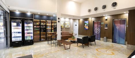 Een hotelhal met een receptiebalie, zithoek, automaten en een digitaal informatiebord.