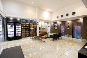 Un hall de hotel con mostrador de recepción, zona de estar, máquinas expendedoras y un panel informativo digital.