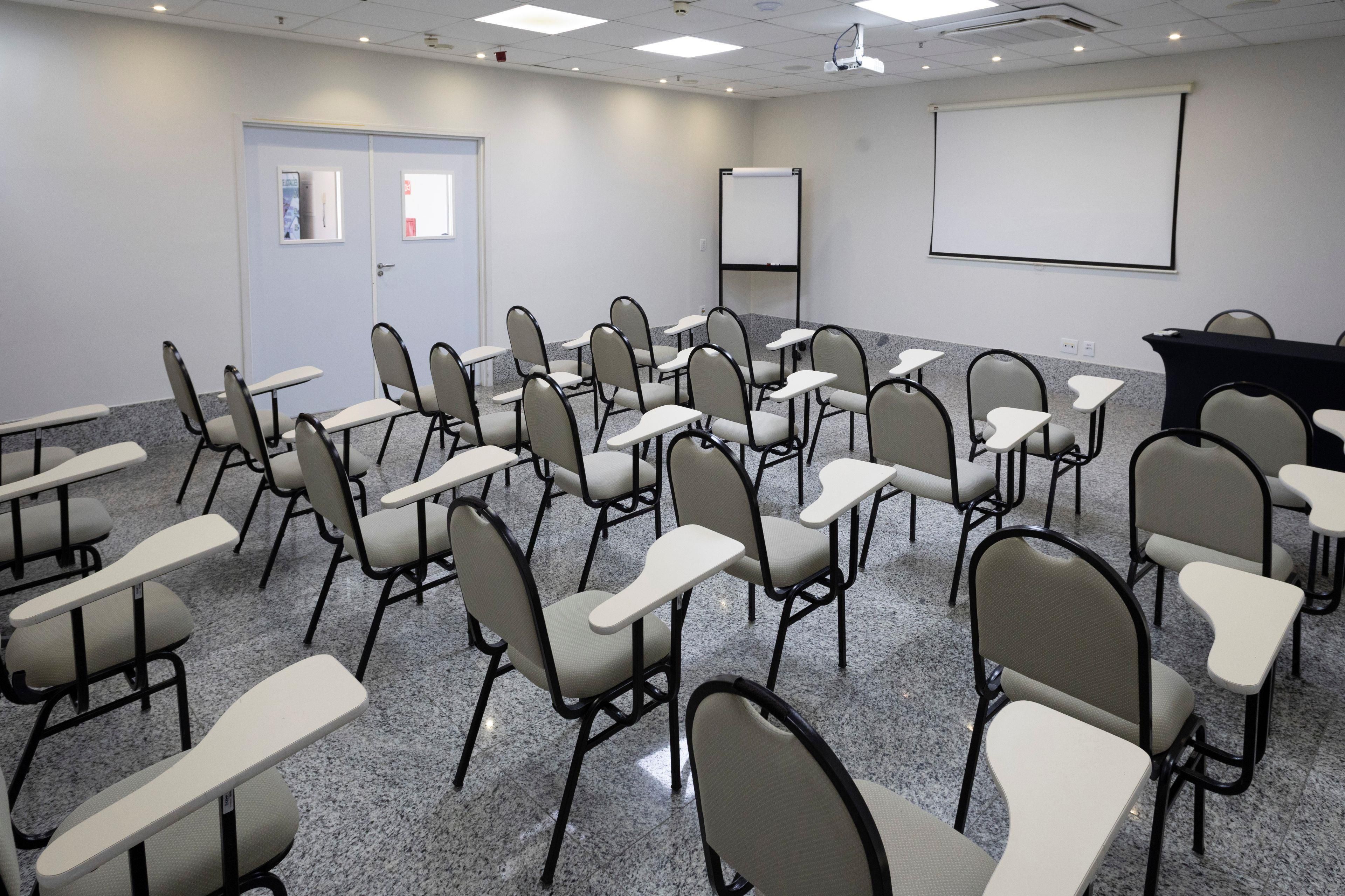 Un salón de conferencias con filas de sillas y mesas, una pantalla de proyector y una pizarra.