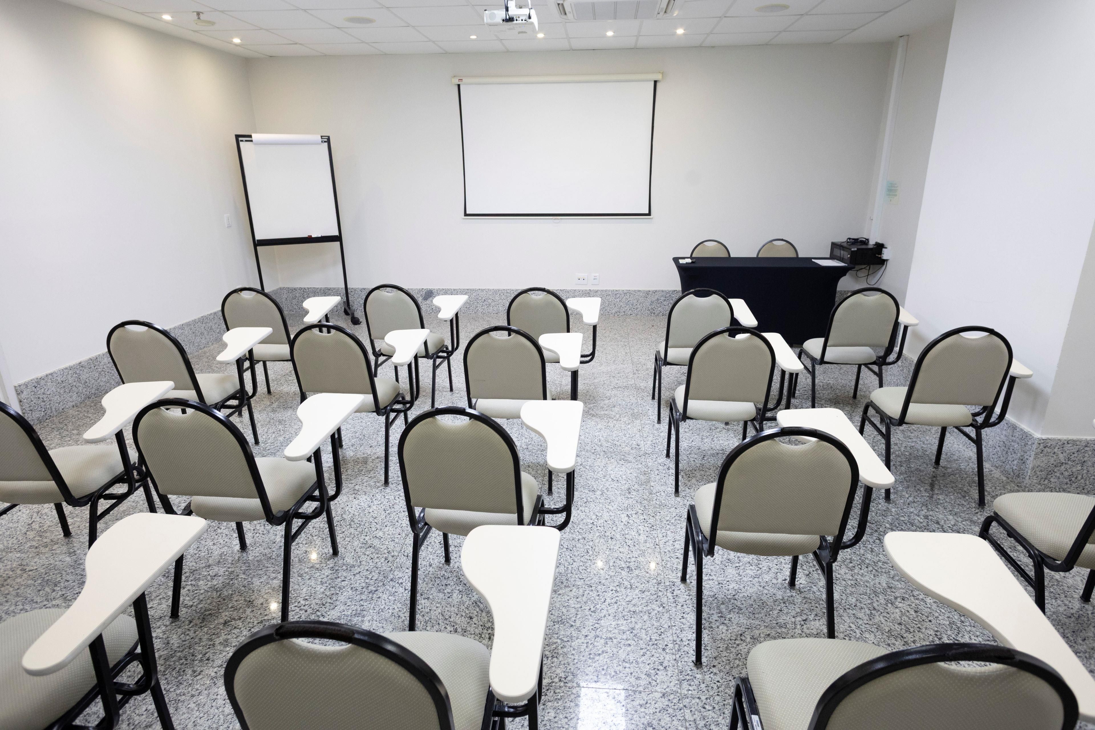 Un salón de conferencias con filas de sillas y mesas, una pantalla de proyector y un atril.