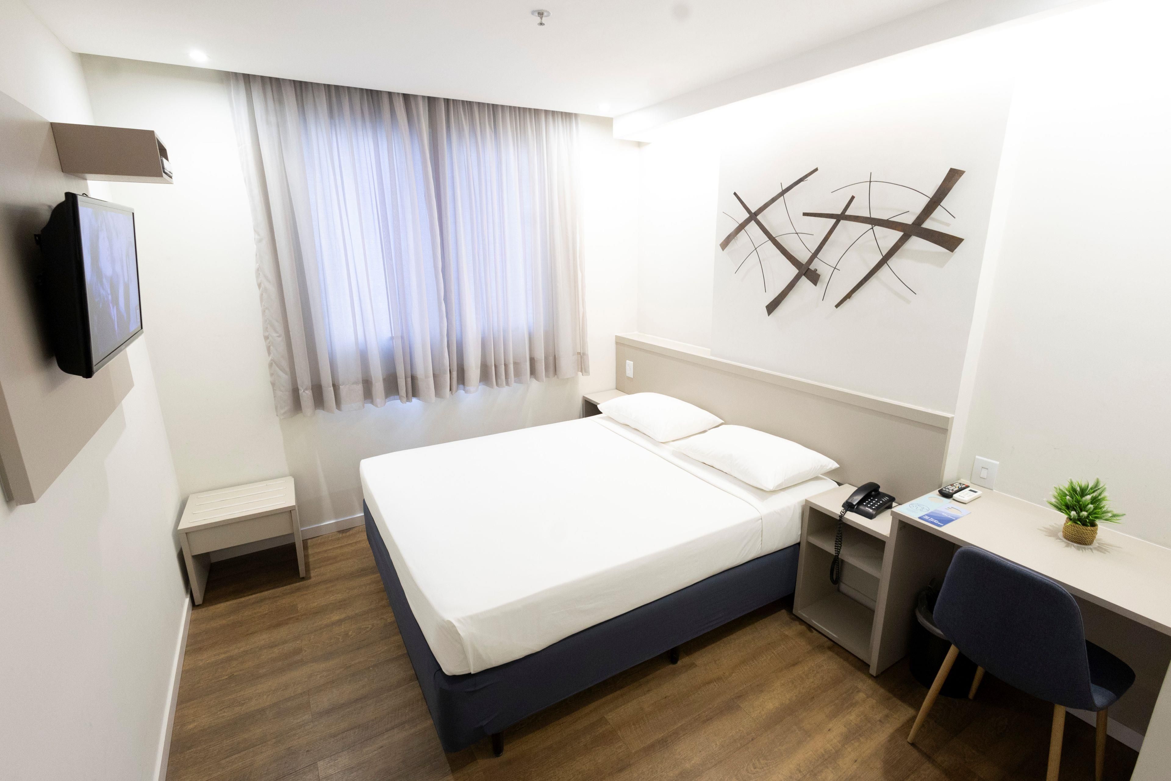 Habitación de hotel con cama, escritorio, silla, televisor, ventana con cortinas y una obra de arte colgada en la pared.