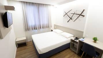 Habitación de hotel con cama, escritorio, silla, televisor, ventana con cortinas y una obra de arte colgada en la pared.