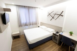 Habitación de hotel con cama, escritorio, silla, televisor, ventana con cortinas y una obra de arte colgada en la pared.