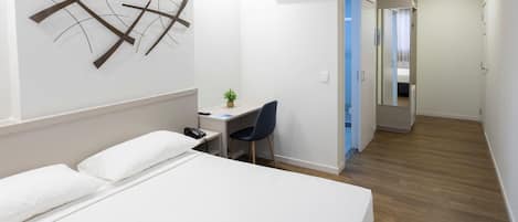 A hotel room with a bed, a desk, a chair, an air conditioner, and a small plant.