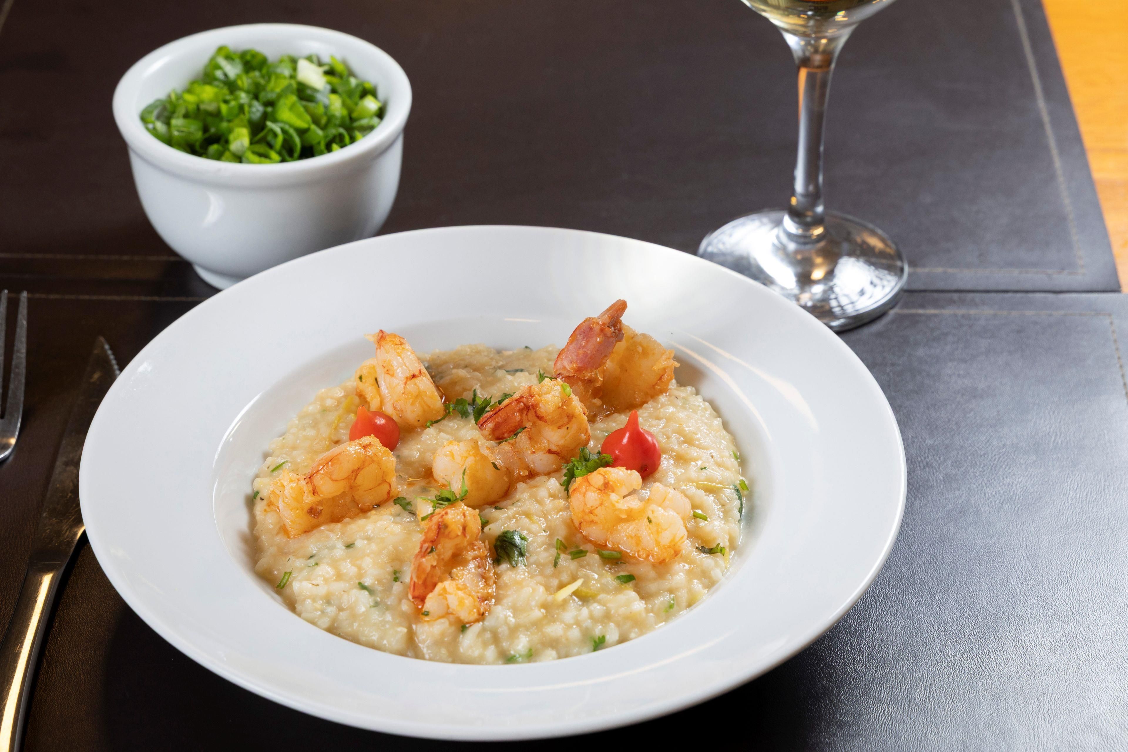Un plato de risotto de camarones con decoración, una copa de vino blanco y un bol de verduras verdes sobre una mesa oscura.