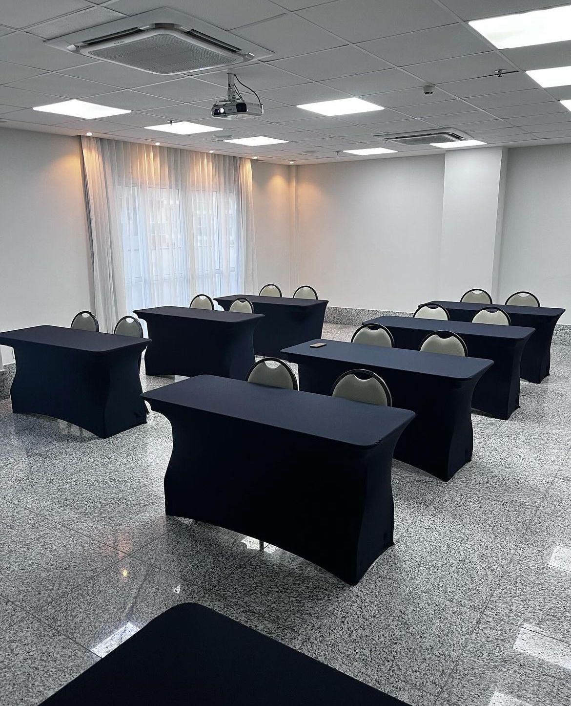 Uma sala de conferências com várias mesas e cadeiras, um projetor e uma grande janela com cortinas.