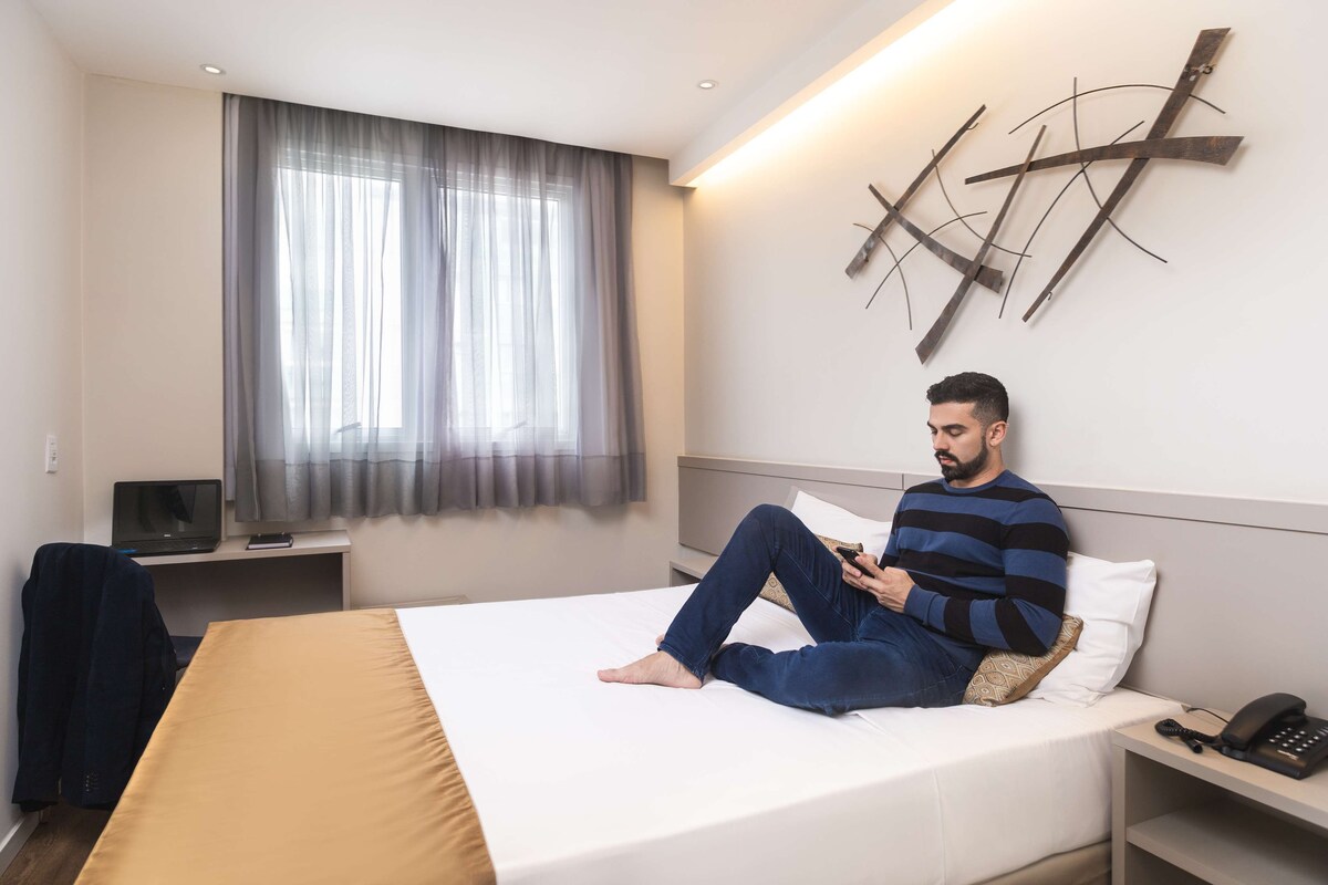 A hotel room with a bed, a desk with a computer, a phone, and a wall-mounted abstract art piece.