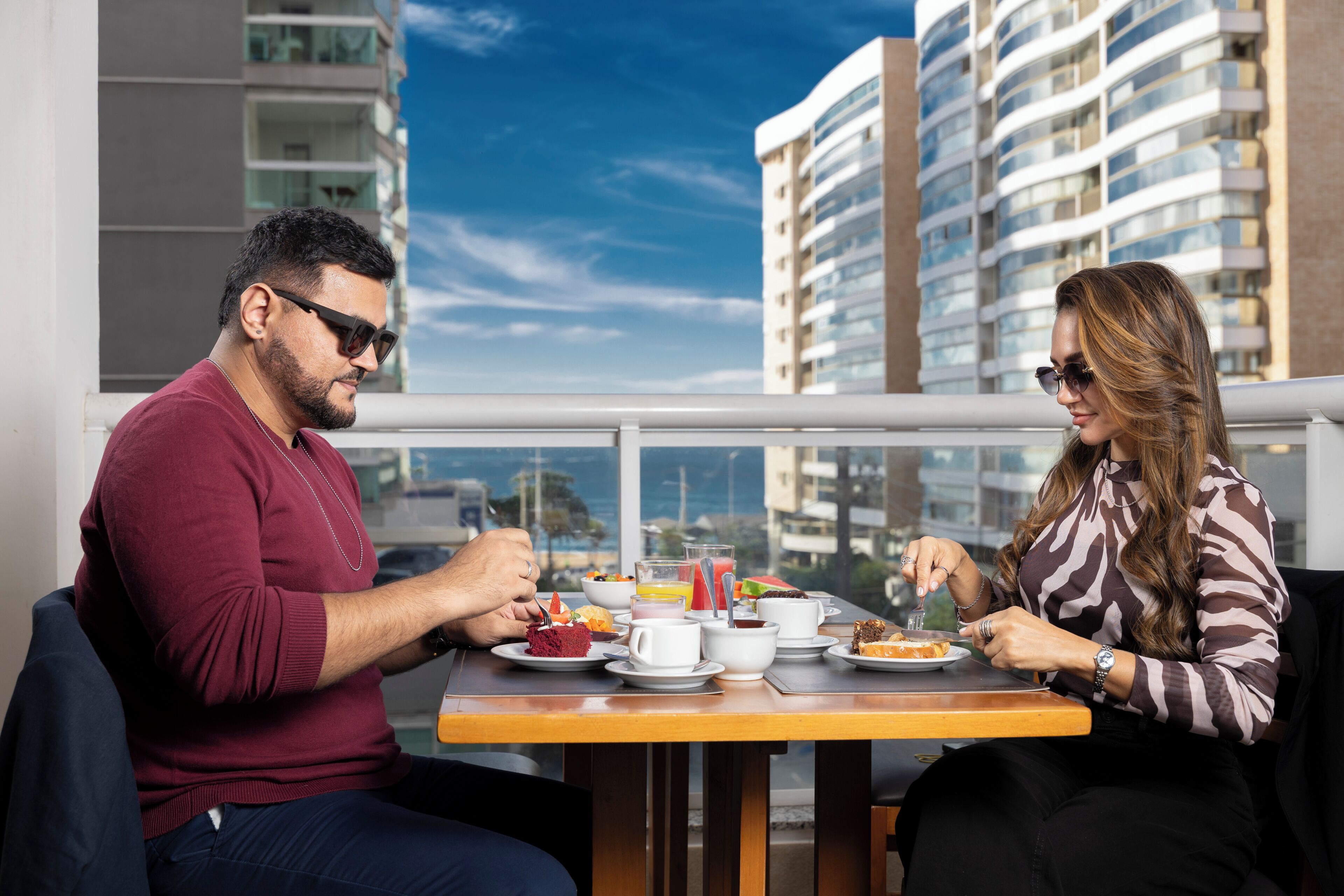 A dining table with a meal, two people eating, and a cityscape view.