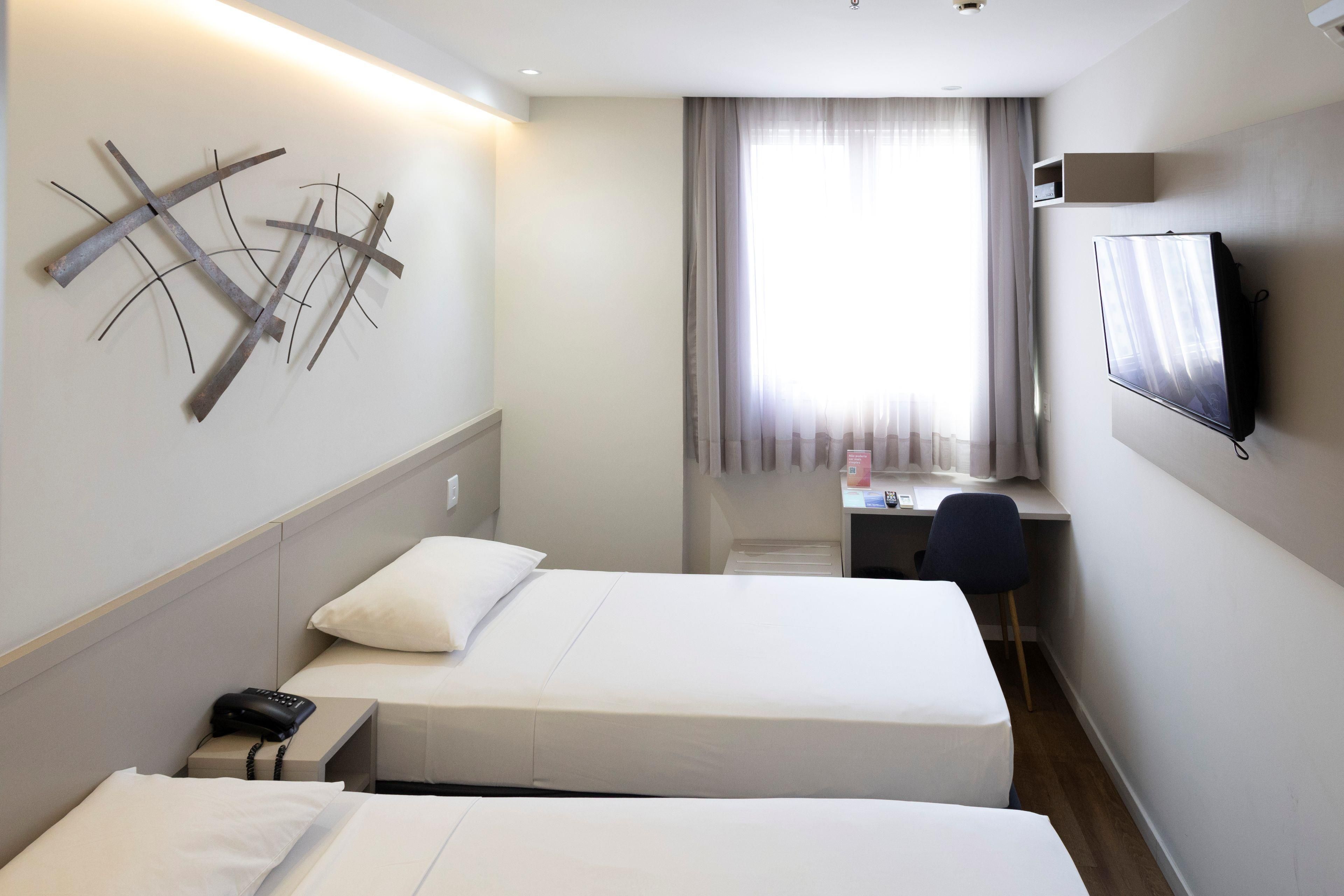 Quarto de hotel com duas camas, uma escrivaninha, uma cadeira, uma televisão e uma peça de arte decorativa na parede.