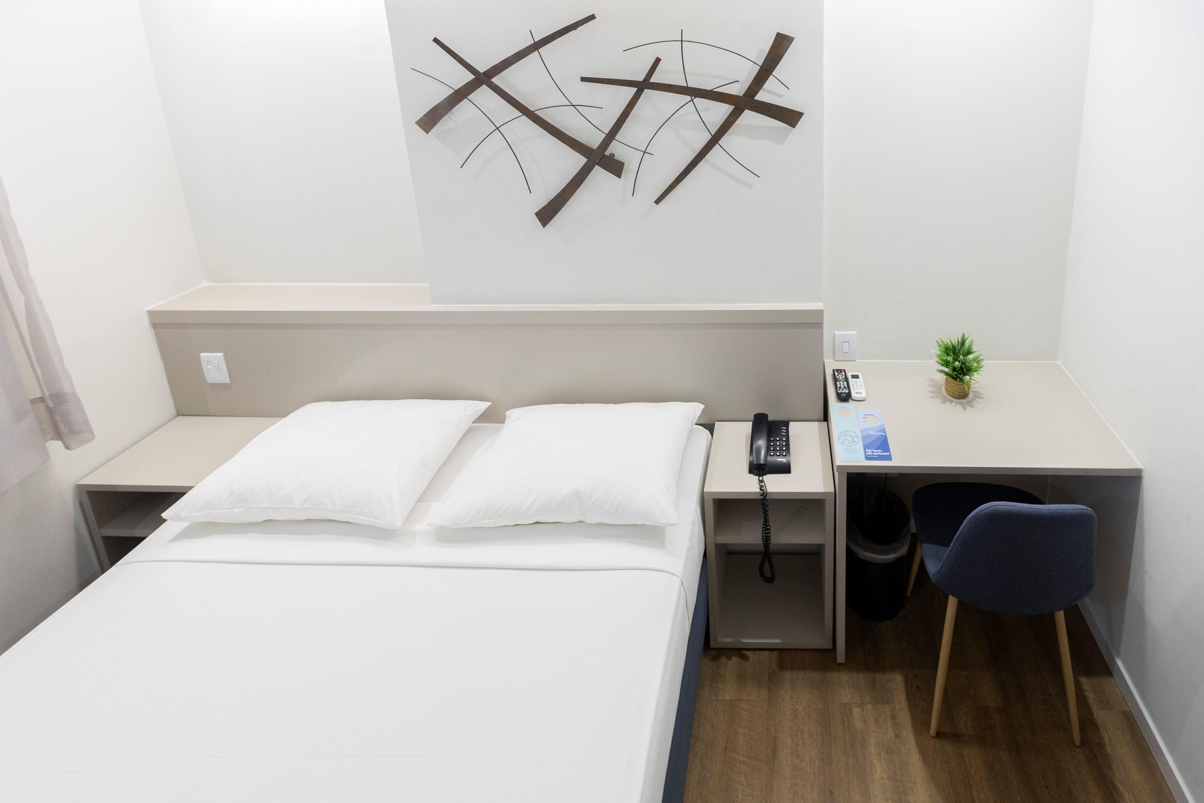 A hotel room with a bed, bedside table, desk, chair, and a wall-mounted telephone.