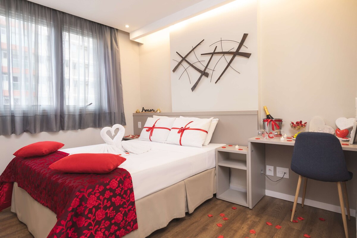 A hotel room with a bed, a desk, and a chair. There are red pillows on the bed and a bouquet of flowers on the desk.