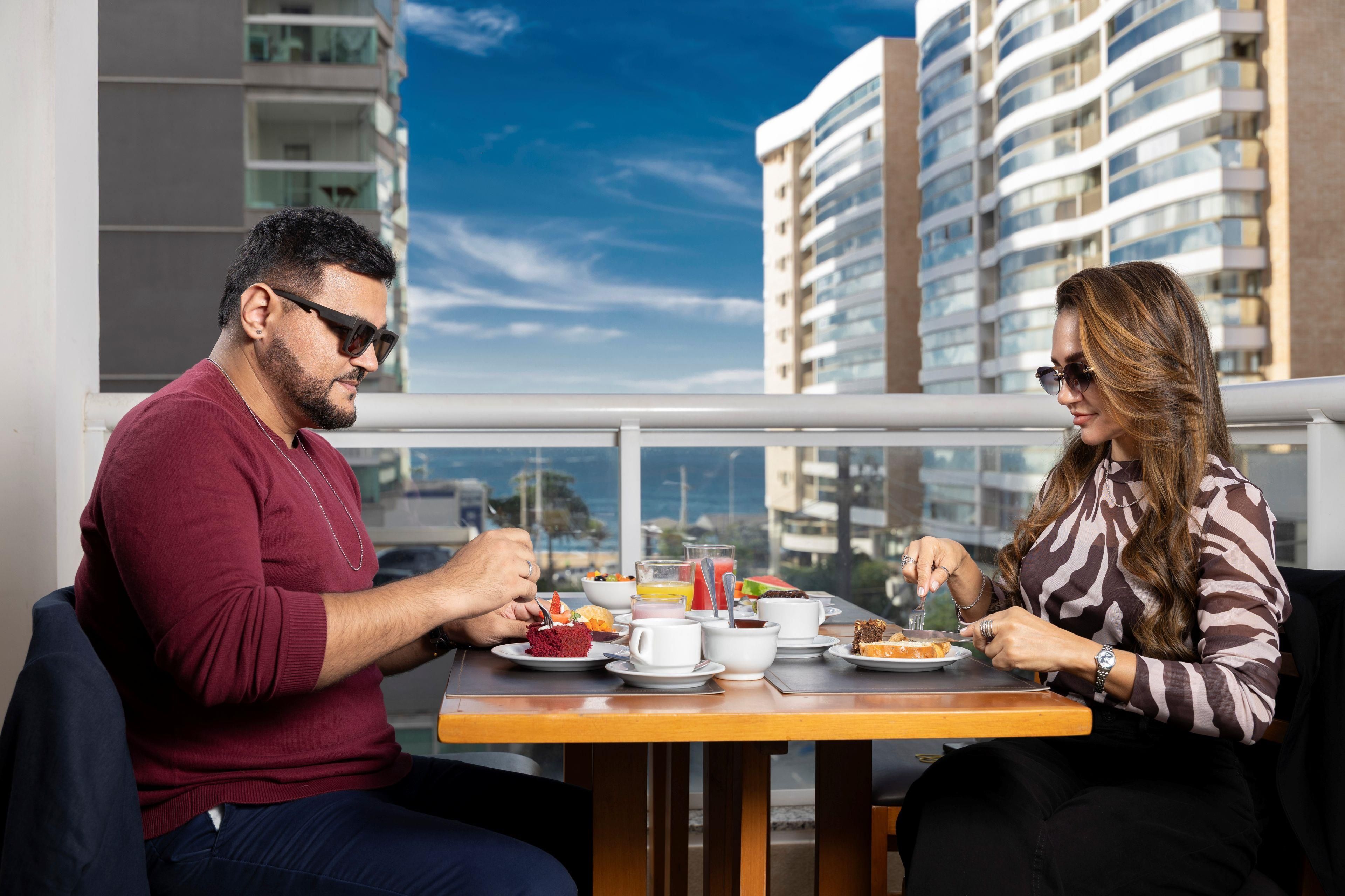Una coppia cena a un tavolo con vista sulla città, un balcone e moderni edifici sullo sfondo.