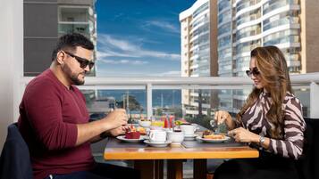 Una coppia cena a un tavolo con vista sulla città, un balcone e moderni edifici sullo sfondo.