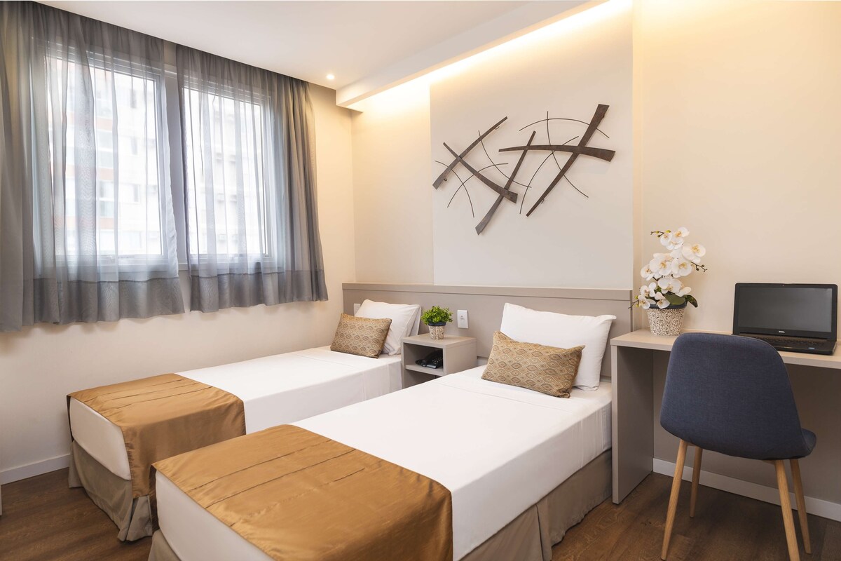 A hotel room with two beds, a desk, a chair, a TV, and a wall-mounted plant.