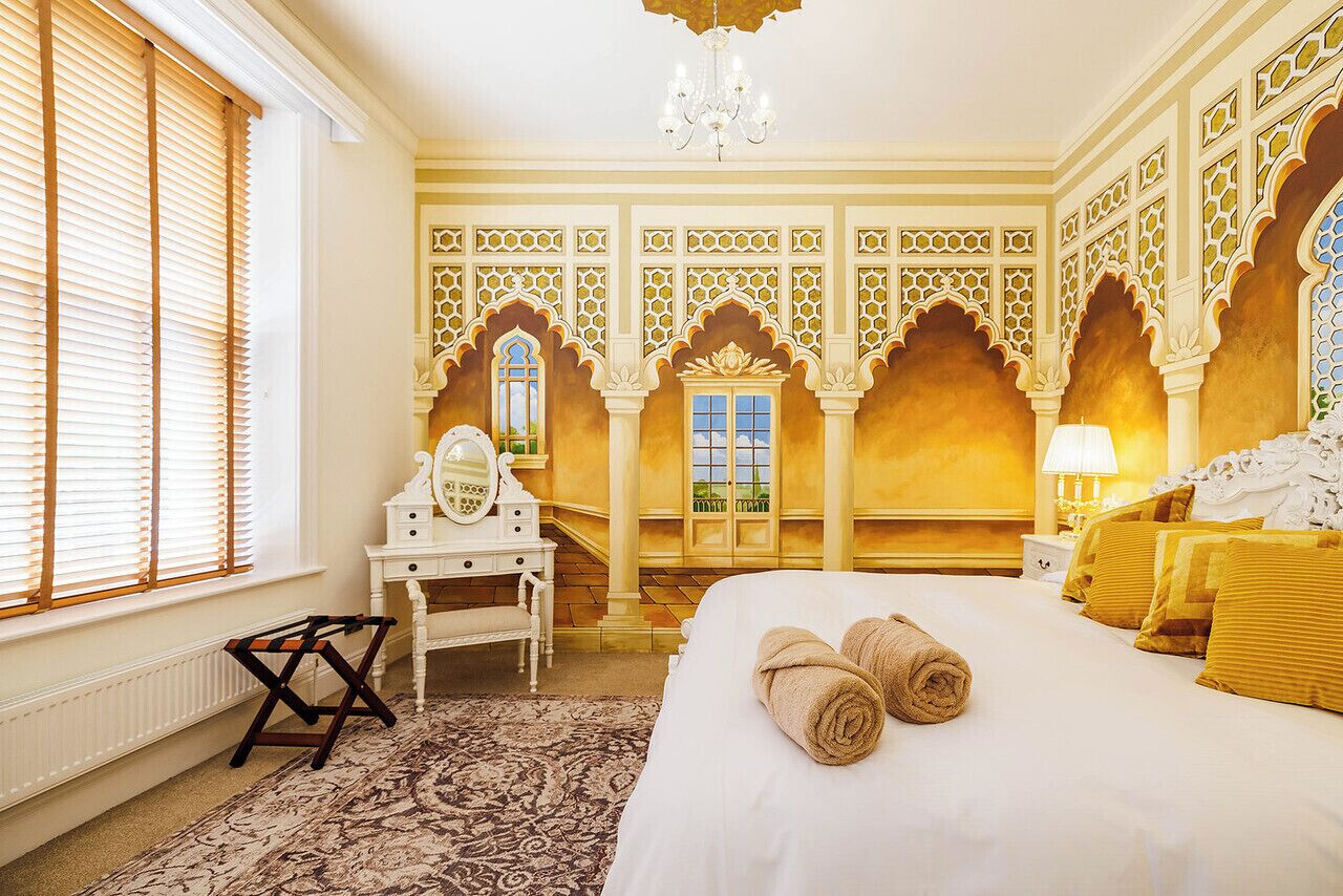 A hotel room with a bed, a vanity with a mirror, a window with blinds, and a chandelier.
