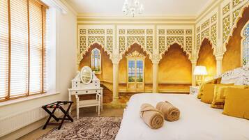 A hotel room with a bed, a vanity with a mirror, a window with blinds, and a chandelier.