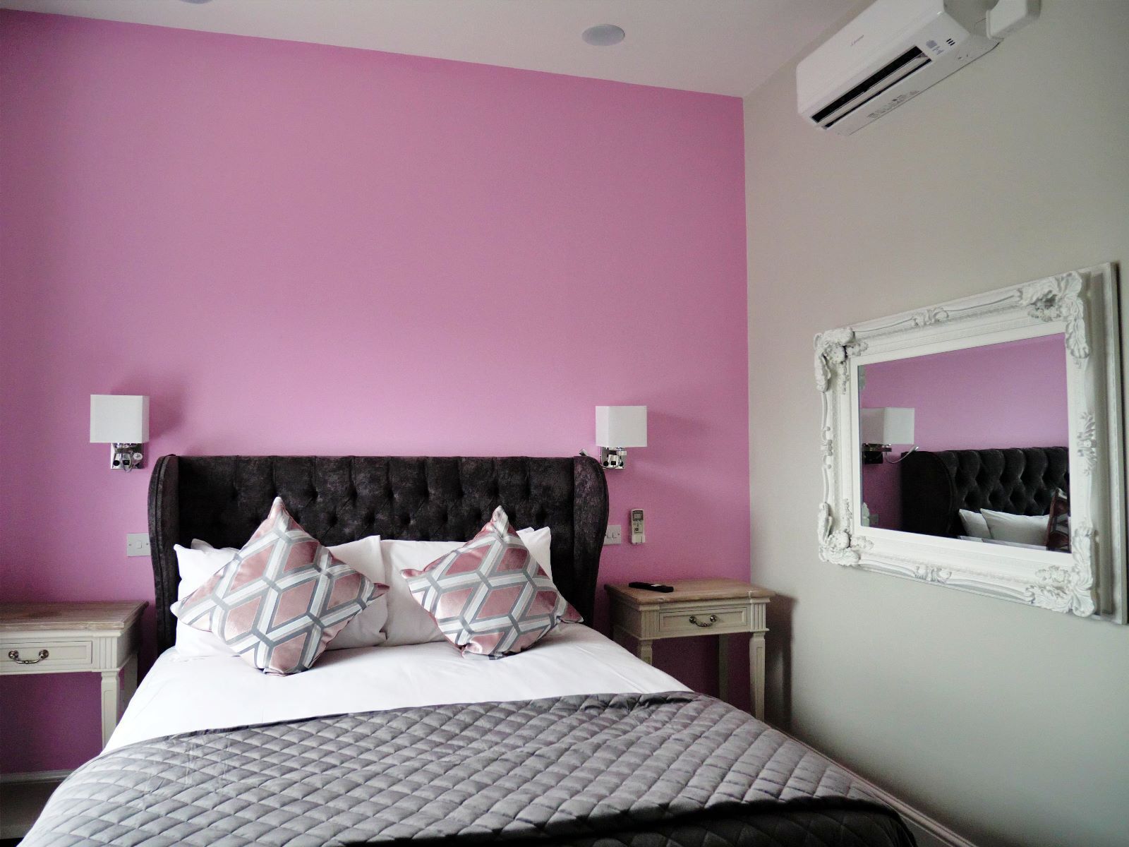 A bedroom with a pink wall, a tufted headboard, two bedside tables with lamps, a mirror, and a bed with patterned pillows.