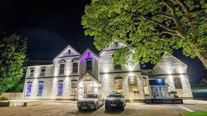Un edificio grande de varios pisos con fachada de piedra, dos coches estacionados frente a él y una entrada bien iluminada.