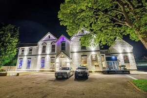 Un edificio grande de varios pisos con fachada de piedra, dos coches estacionados frente a él y una entrada bien iluminada.