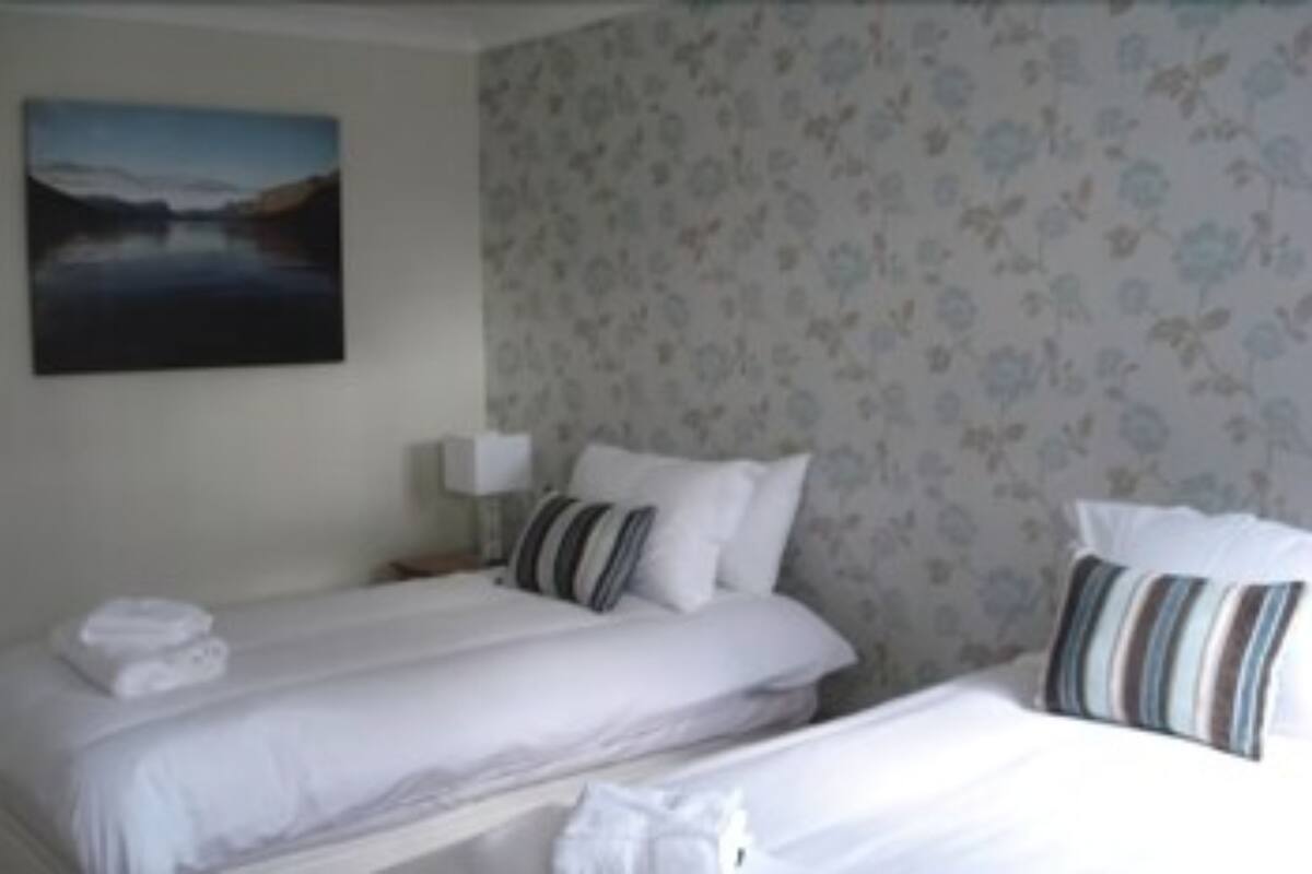 Une chambre d’hôtel avec deux lits, un papier peint à motifs floraux, un tableau au mur et une table de chevet avec une lampe.