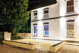 Un edificio blanco con fachada de ladrillo, varias ventanas y un acceso pavimentado con macetas.