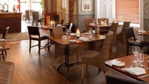 Un comedor con mesas y sillas de madera, un candelabro y vistas de la cocina a través de un vano abierto.