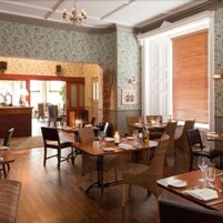 Un comedor con mesas y sillas de madera, un candelabro y vistas de la cocina a través de un vano abierto.