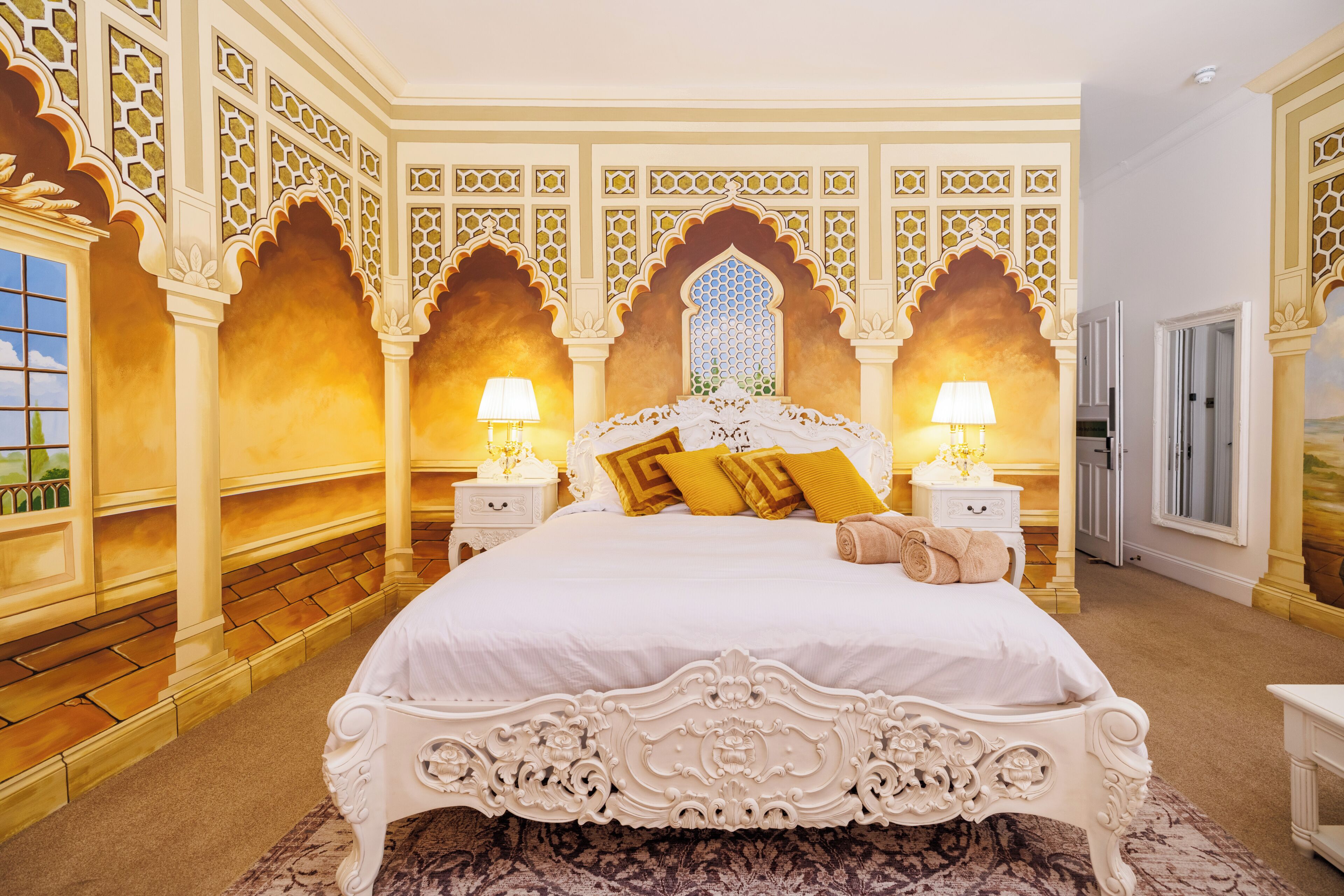 A large bed with ornate headboard, two bedside tables with lamps, and a patterned rug.