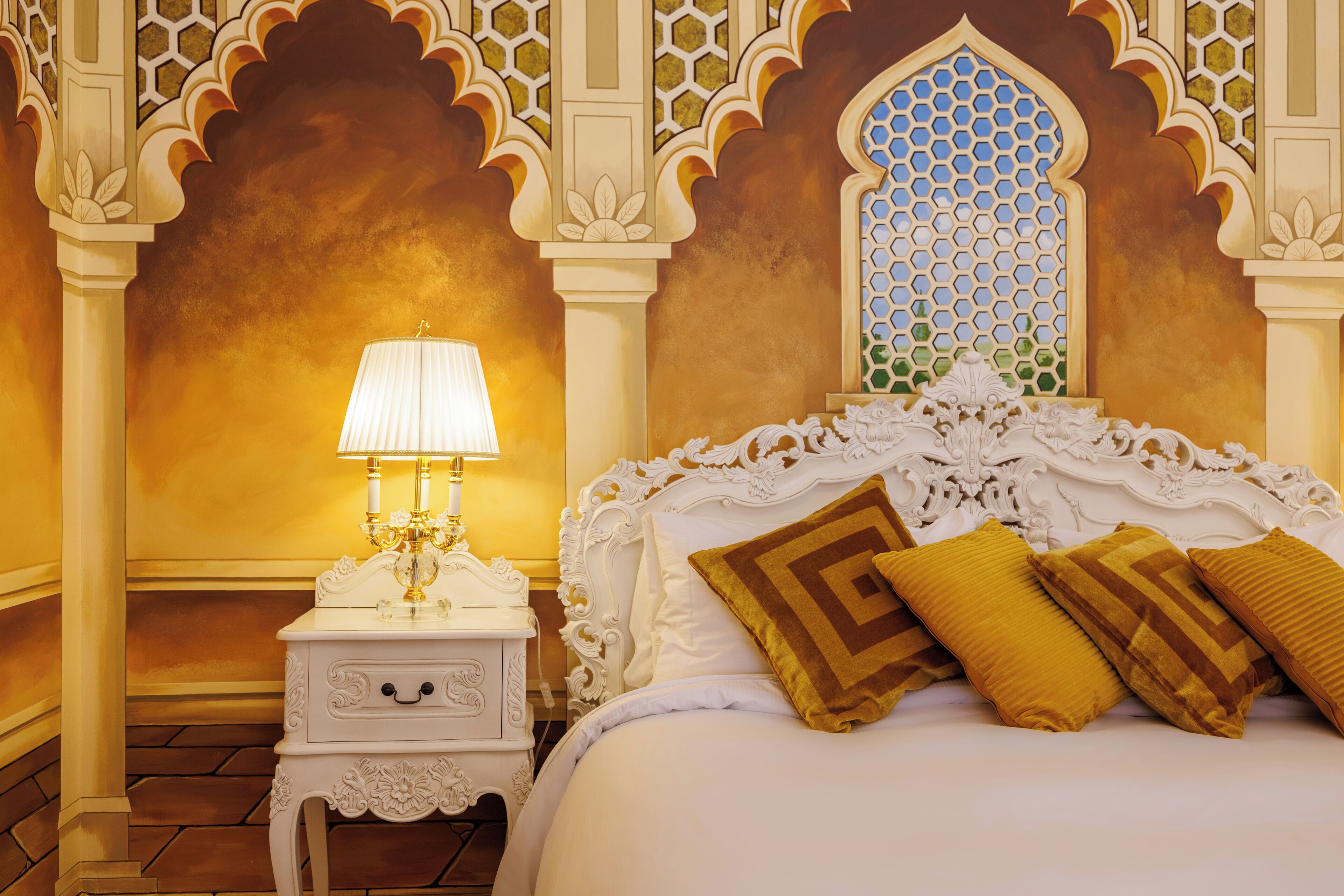 A bedroom with a white ornate bed, a nightstand with a lamp, and a decorative wall with a geometric window.