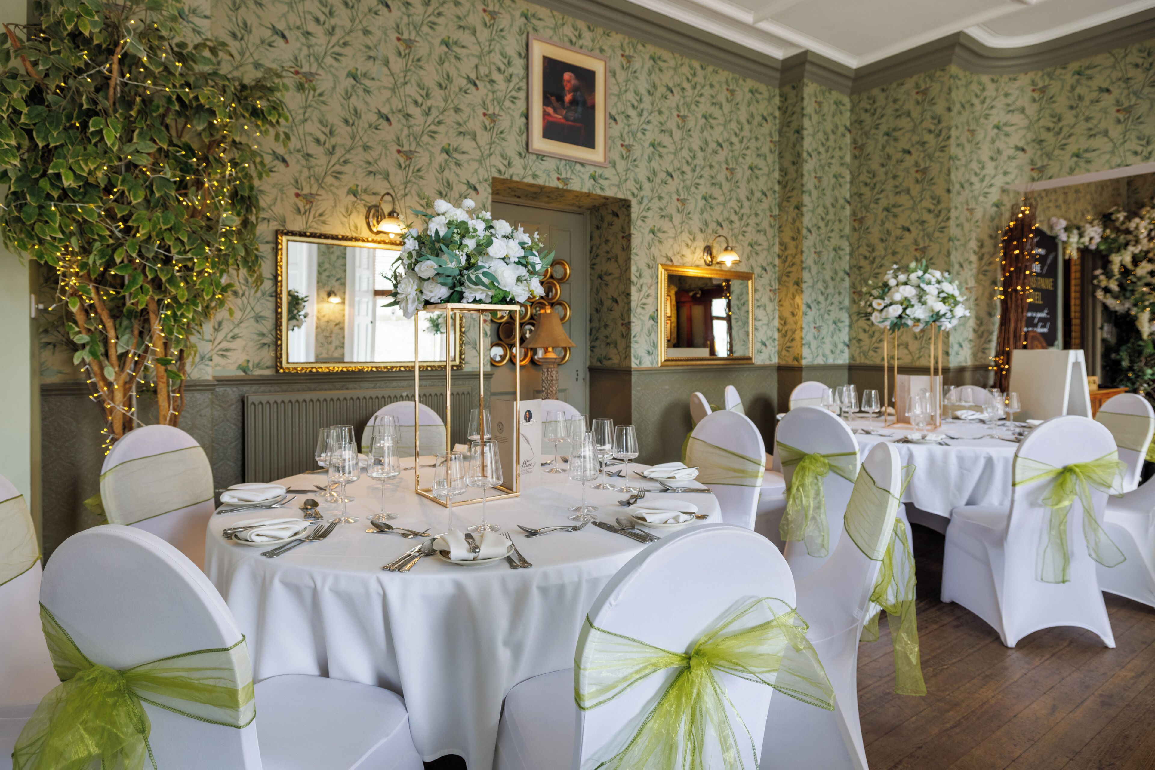 Een formele eetzaal met ronde tafels, witte stoelen met groene linten, bloemstukken en spiegels aan de muur.