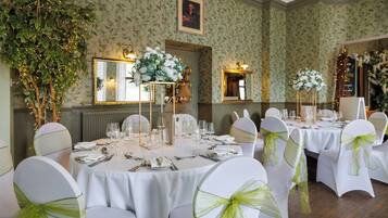 Een formele eetzaal met ronde tafels, witte stoelen met groene linten, bloemstukken en spiegels aan de muur.