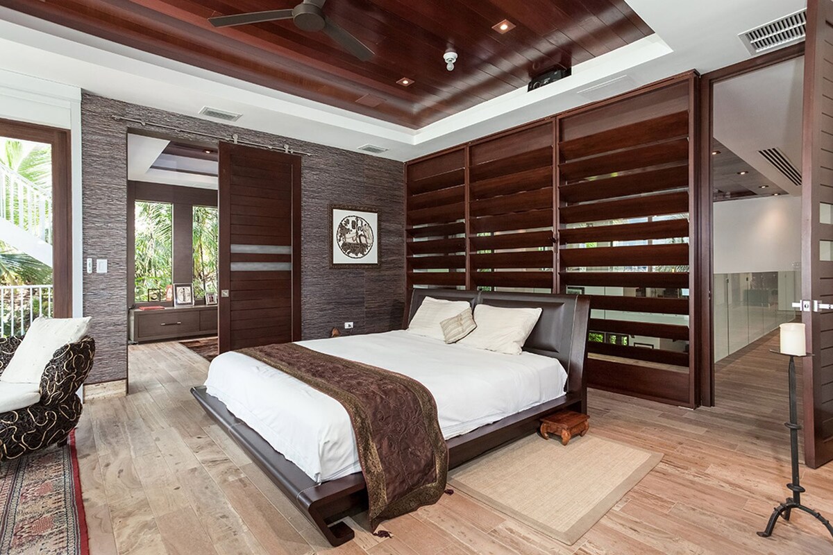 A modern bedroom with a large bed, a wooden headboard, a ceiling fan, and a view of the outdoors.
