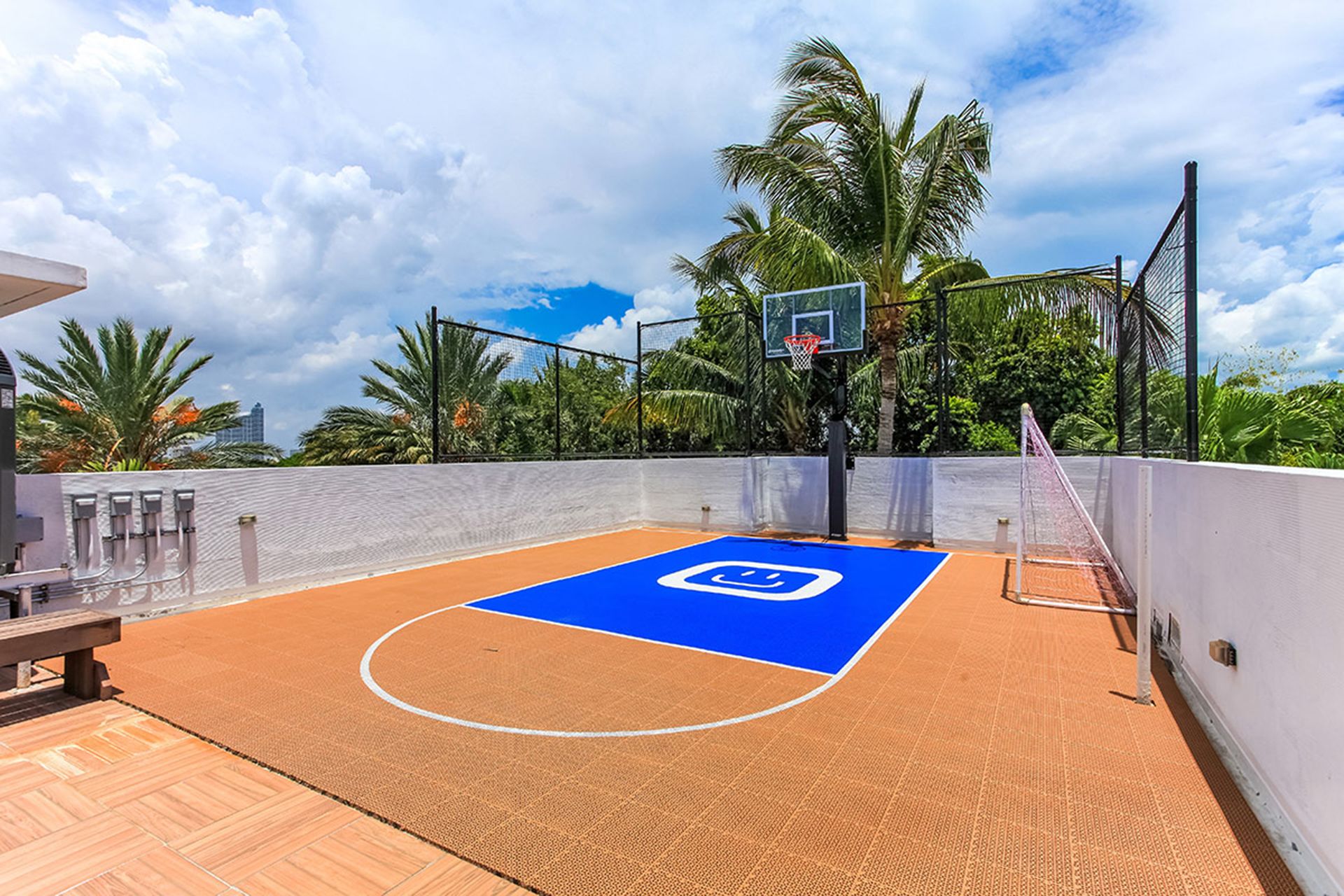 An outdoor basketball court with a blue center circle, surrounded by a white wall and palm trees.