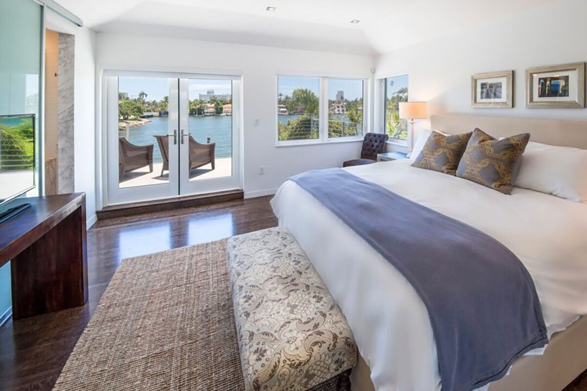 A spacious bedroom with a large bed, a bench, and a view of a body of water through sliding glass doors.