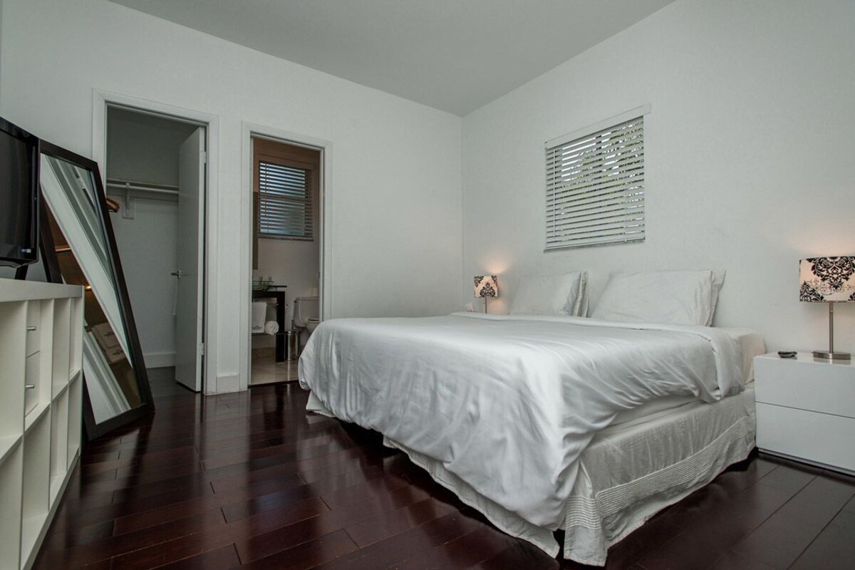 A bedroom with a large bed, a bedside table, a mirror, and a window with blinds.