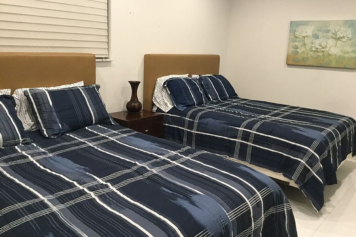 A hotel room with two beds, each with a headboard and a matching pillow. There is a nightstand between the beds with a vase on it.