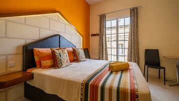 Una habitación de hotel con una cama grande, un cabecero con diseño de ladrillo, una ventana con cortinas estampadas y una pequeña mesa con una silla.