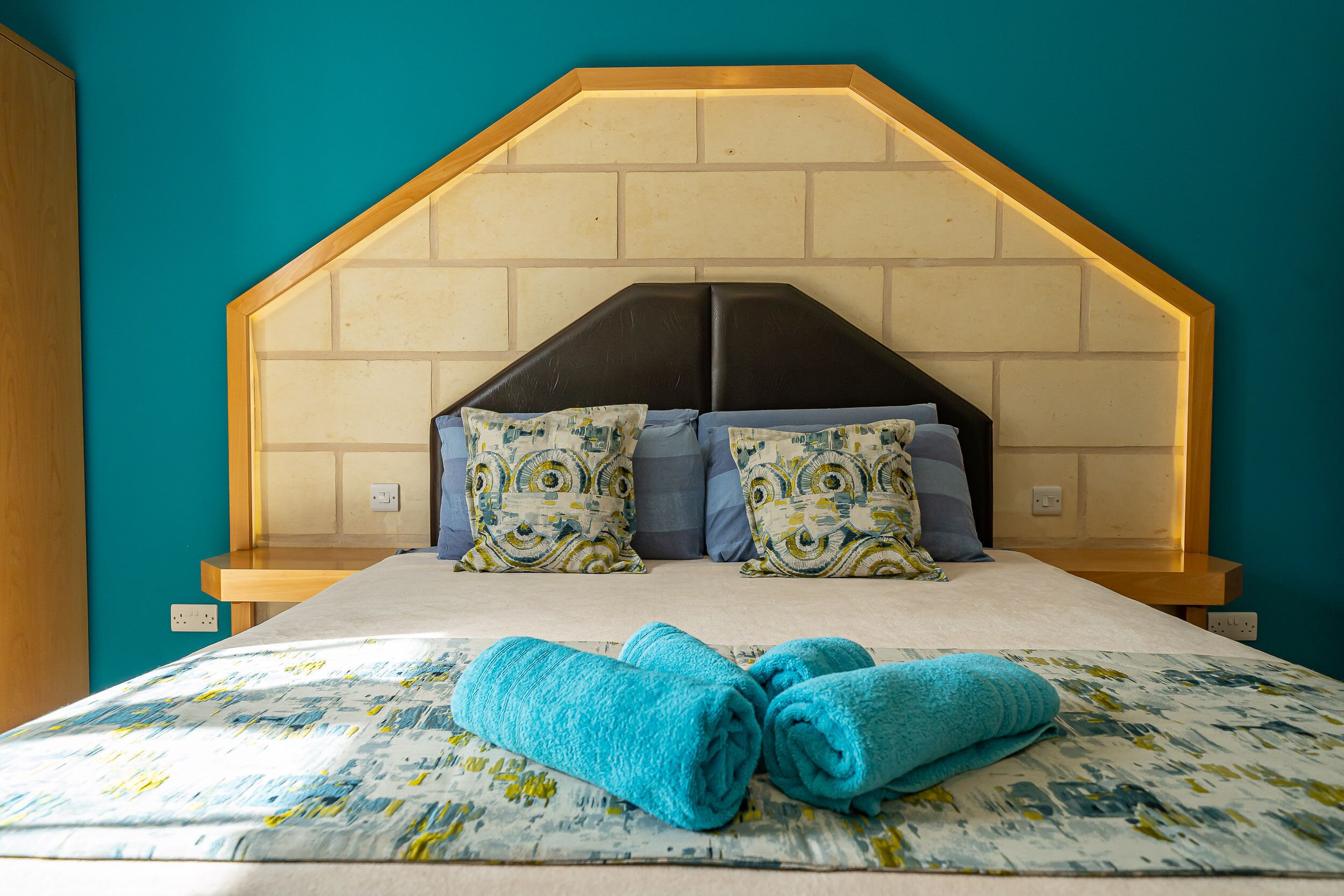 A bedroom with a wooden headboard, patterned pillows, and rolled towels on the bed.