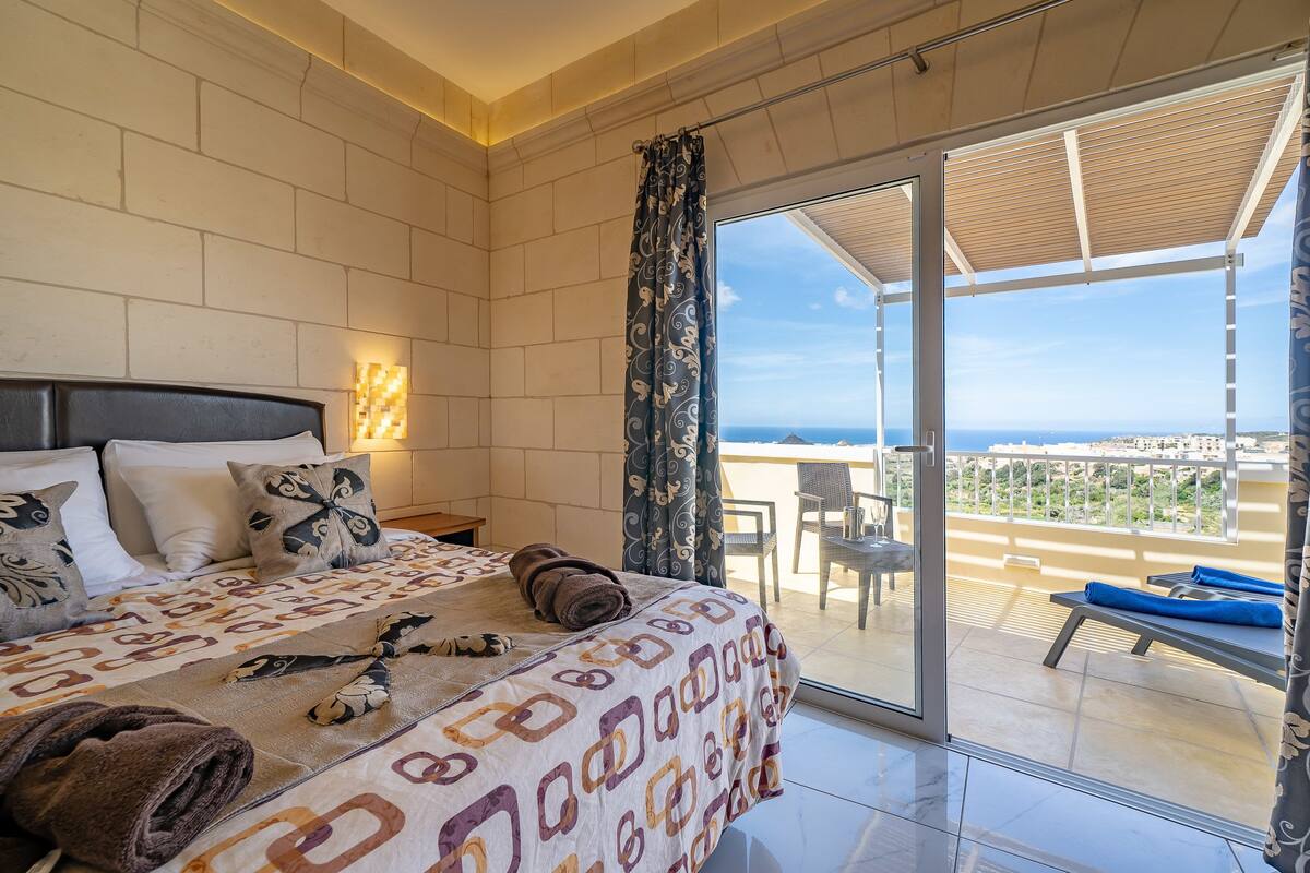 A bedroom with a bed, pillows, a blanket, and a view of the balcony and outside.
