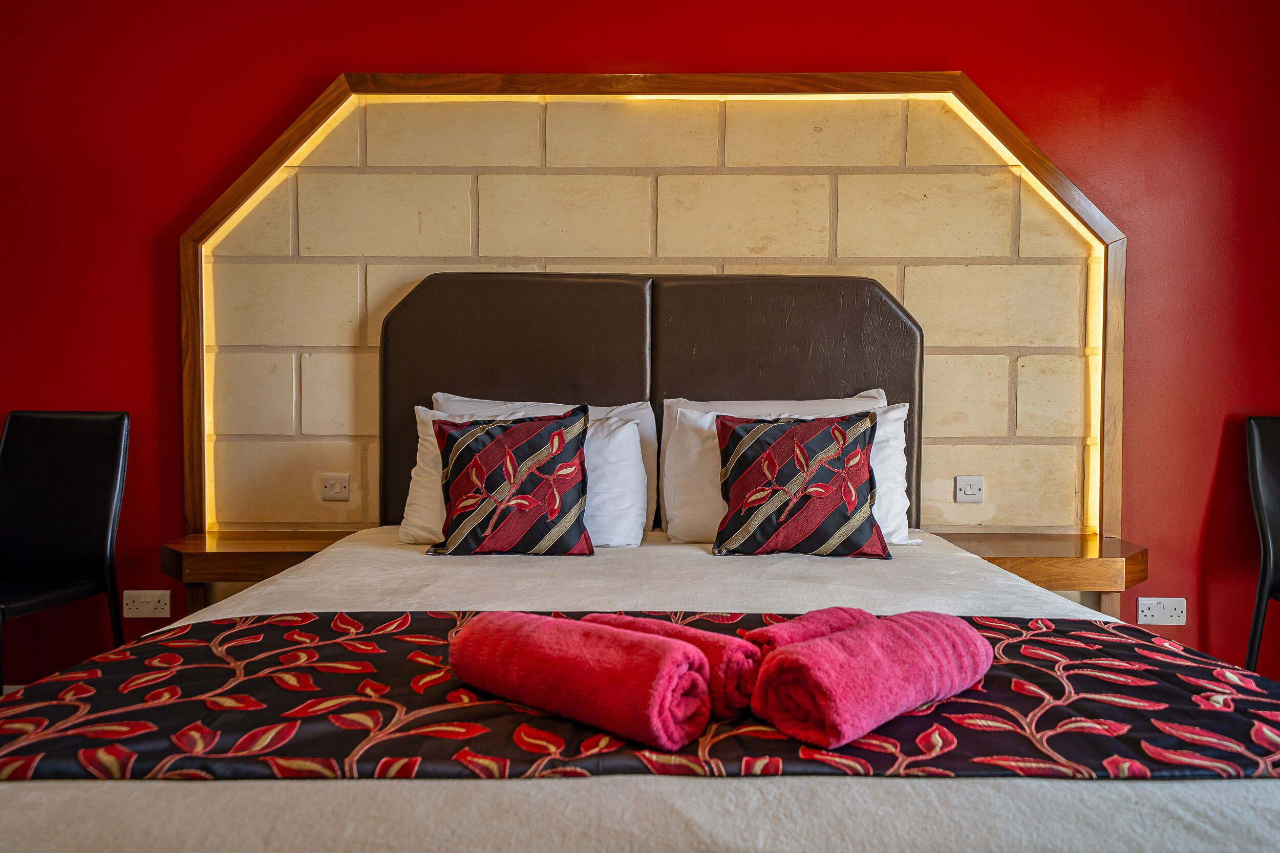 A hotel room with a brick-patterned headboard, two pillows with a red and black pattern, and a bed with a red and black floral bedspread.