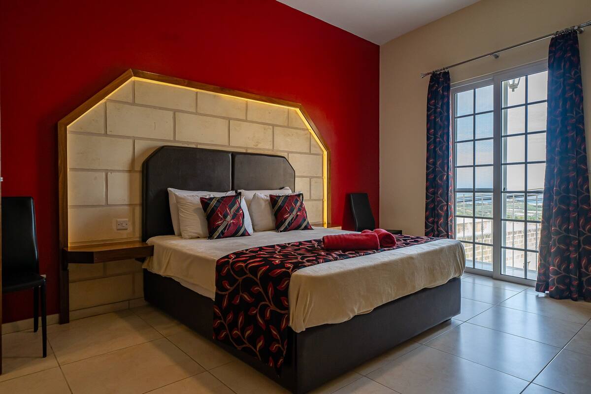 A bedroom with a large bed, a headboard with built-in lighting, a window with curtains, and a red accent wall.