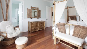 Een slaapkamer met een hemelbed, een rieten stoel, een houten dressoir en een plafondventilator.