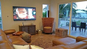 A living room with a large flat-screen TV, a brown leather sofa, a wicker chair, and a small cabinet. - 6 Bedroom Homes in Miami by TMG (Miami)